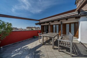 A rooftop patio with a wooden table and chairs, a red wall, and a view of buildings in the background. - GuestHost - Cozy apartment of 143 sqm, able to accommodate up to 5 people, located on the third floor of a building with lift in the heart of Udine. The location is great! In about 10 minutes walk you can reach Via Mercatovecchio, Piazza della Libertà a (Udine)