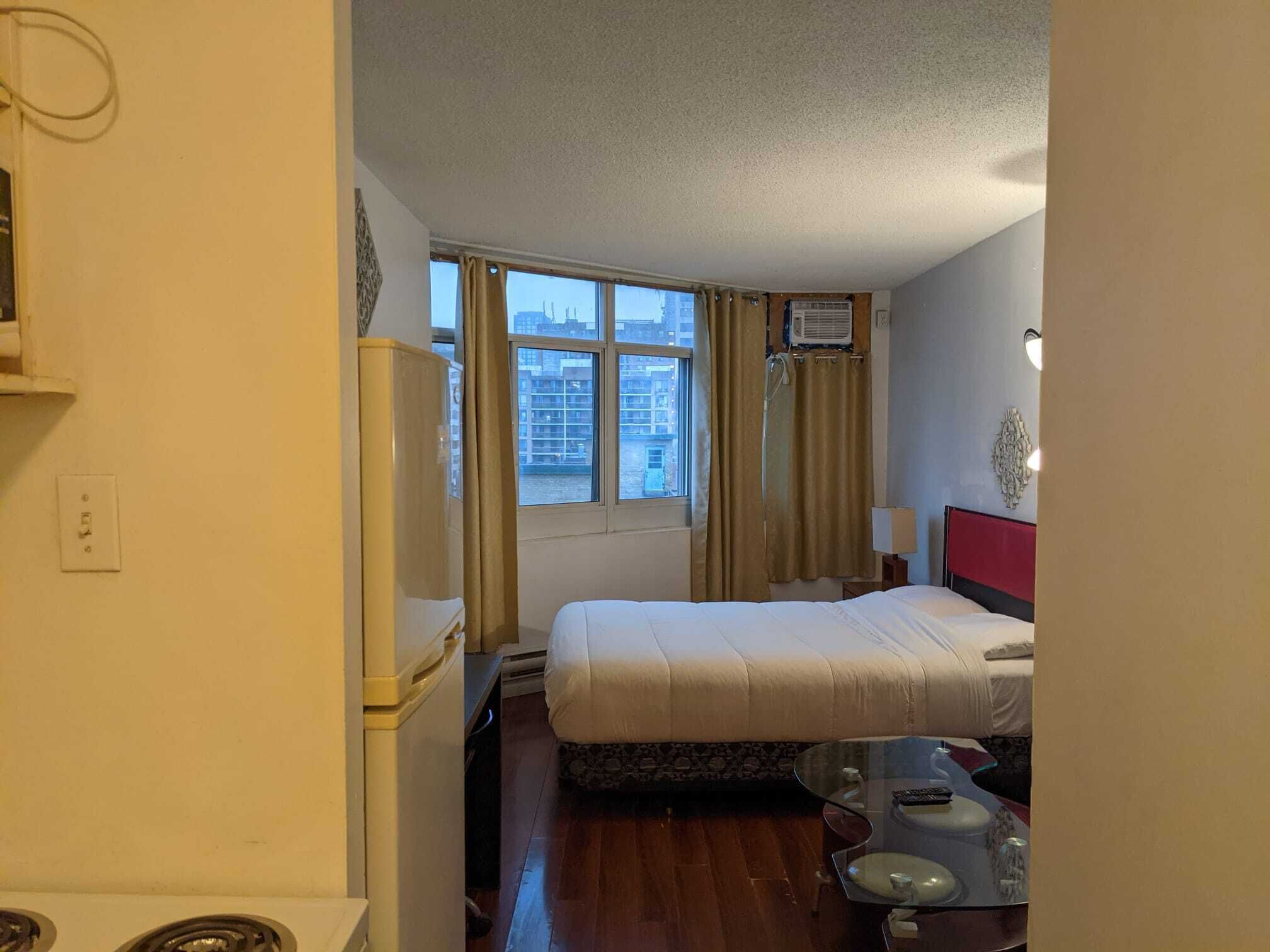Een hotelkamer met een bed, een tafeltje, een stoel, een raam met gordijnen en een wandairconditioner.