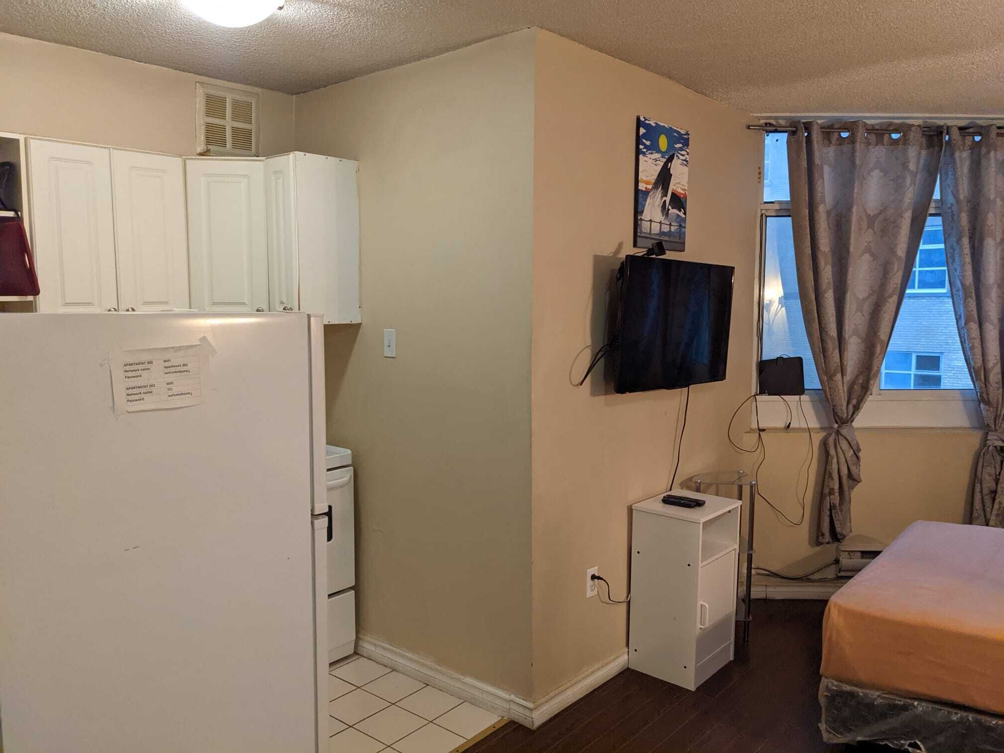 A small, compact room with a white refrigerator, a small white cabinet, a television mounted on the wall, and a window with curtains.