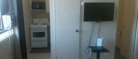 A kitchen with a stove, oven, and microwave. There is a door leading to another room, a television mounted on the wall, and a small table with a power strip.