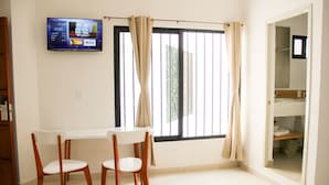 Una habitación con una mesa de comedor y sillas, un televisor instalado en la pared y una ventana con cortinas.