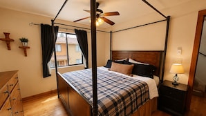 Un lit à baldaquin avec une couvre-lit à carreaux, une table de chevet en bois avec une lampe, un ventilateur de plafond et une fenêtre avec des rideaux.