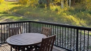 Une terrasse dotée d’une table ronde et de chaises, entourée d’une rambarde noire, et offrant une vue sur une zone boisée aux couleurs automnales.