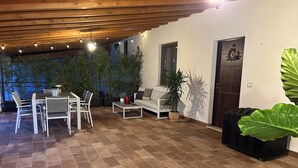 Een terras buiten met een houten plafond, betegelde vloer, witte meubels en potplanten.