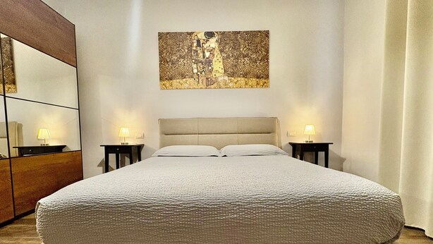 Een net opgemaakt bed met een gebobbelde witte dekbedovertrek, twee bedlampjes en een kunstwerk in een lijst erboven.