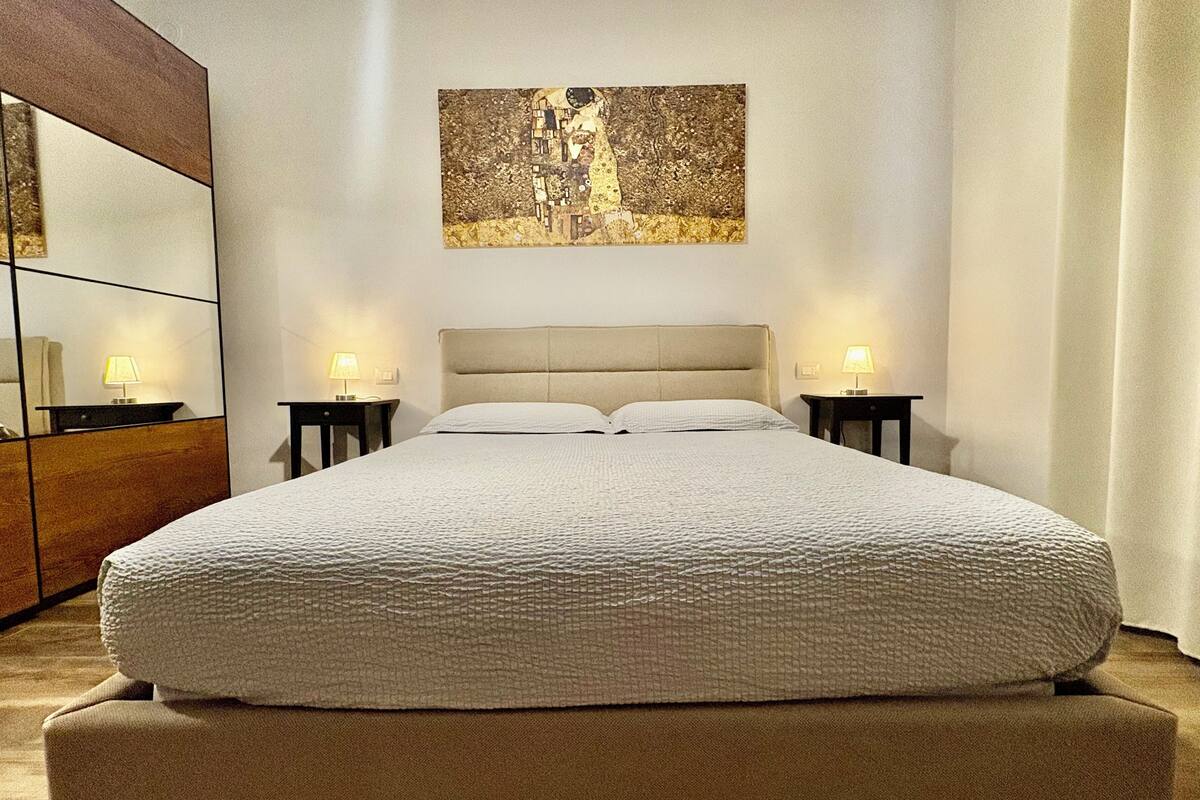 A neatly made bed with a textured white duvet, two bedside lamps, and a framed artwork above the bed.