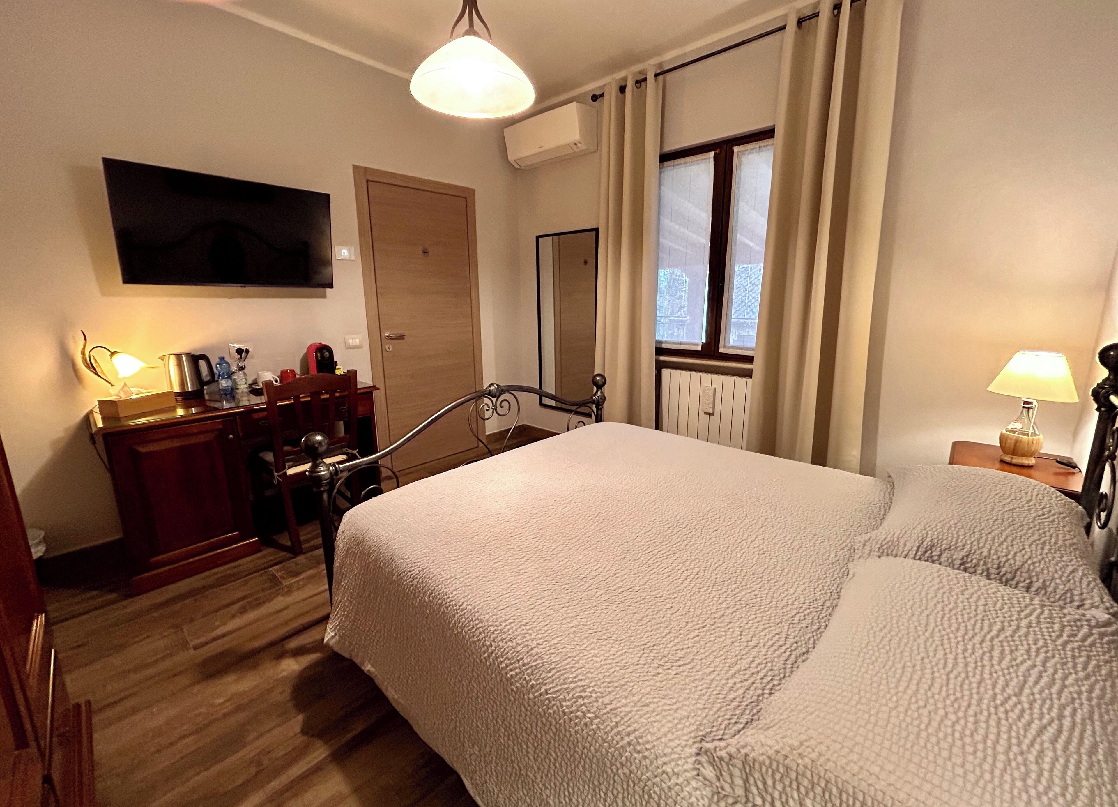 A hotel room with a large bed, a desk with a lamp, a chair, a television on the wall, and a window with curtains.