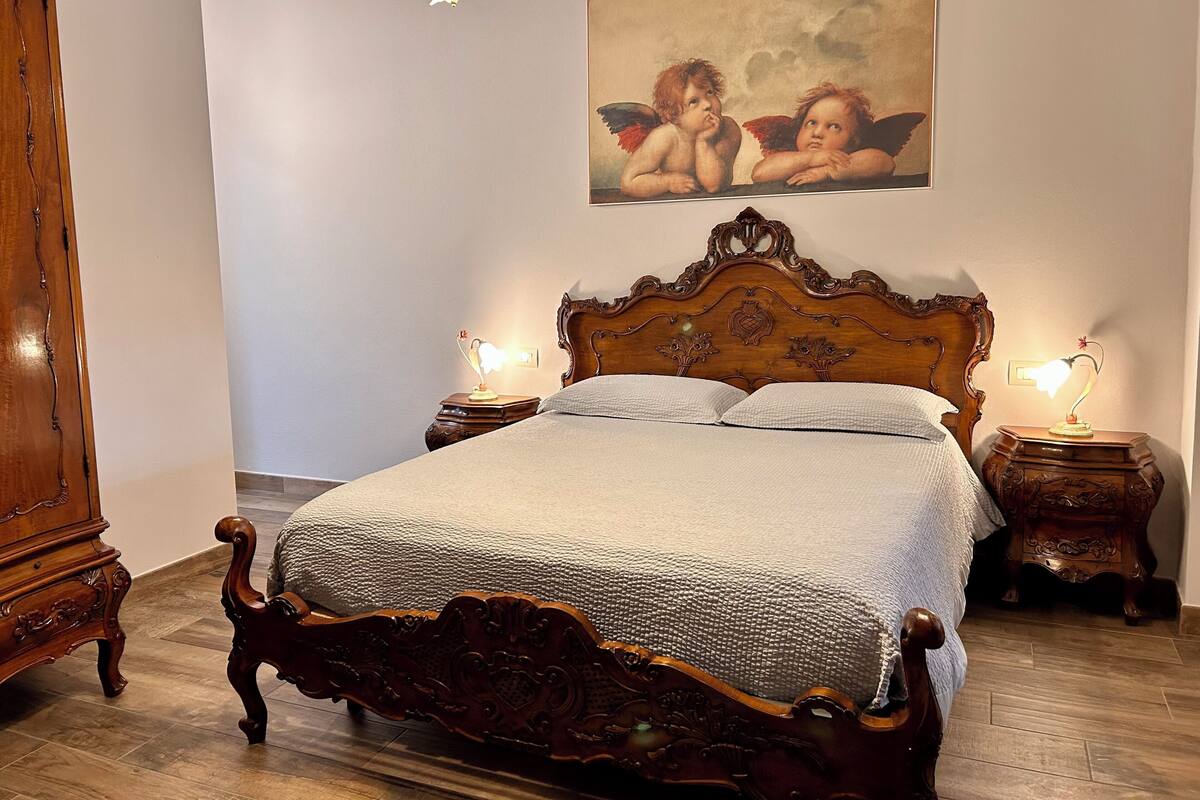 A bedroom with a wooden four-poster bed, two bedside tables with lamps, a wooden wardrobe, and a painting on the wall.