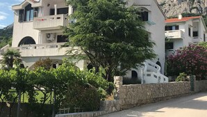 Un edificio moderno de dos pisos con fachada de piedra, varios balcones y un árbol verde frondoso al frente.