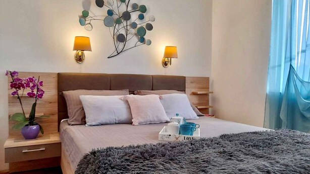Un dormitorio moderno con una cama grande, dos mesitas de noche, un adorno en la pared y una ventana con cortinas translúcidas.