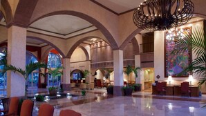 Eine geräumige Lobby mit hohen Decken, rundbogigen Säulen und einem Chandelier. Es gibt mehrere Sitzbereiche mit Stühlen und Tischen, und Blumentöpfe sind überall verteilt.