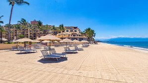 Ein Strand mit Liegestühlen und Sonnenschirmen, ein Hotelgebäude im Hintergrund und Palmen.