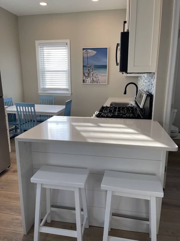 A modern kitchen with a central island, white bar stools, a dining area with blue chairs, and a beach-themed wall art.