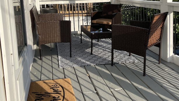 Un espace extérieur aménagé, abrité et doté de meubles en osier et d’un tapis à motifs, un paillasson avec l’inscription « Hourra ! Vous êtes là ! » et une vue sur le quartier.