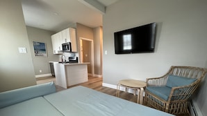 Une chambre d’hôtel moderne avec un grand lit, une petite table, une chaise en osier, un téléviseur et, au fond, une kitchenette.