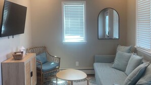 Un salon avec un ventilateur de plafond, un téléviseur fixé au mur, un canapé, un fauteuil, une petite table et un tapis à motifs.