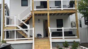 Un immeuble résidentiel de deux étages doté d’une terrasse en bois, d’un escalier et d’une voiture garée devant.