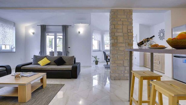 A modern living room with a sofa, coffee table, and a kitchen area with a stone column. - Villa with pool, BBQ and games room (Valbandon)