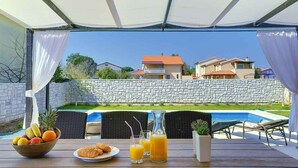 A breakfast setup with a bowl of fruit, croissants, and orange juice on a wooden table under a canopy, overlooking a swimming pool and houses. - Villa with pool, BBQ and games room (Valbandon)