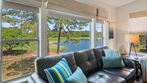 A modern living room with a leather sofa, large windows offering a view of a golf course and lake. - 3BR/2BA Pet-Friendly Condo with Pool on True Blue Golf Course (Pawleys Island)