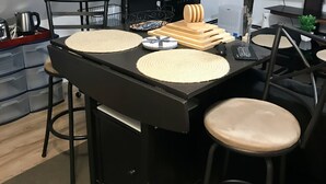 A modern kitchen with a black countertop, two bar stools, a coffee machine, and a TV mounted on the wall.