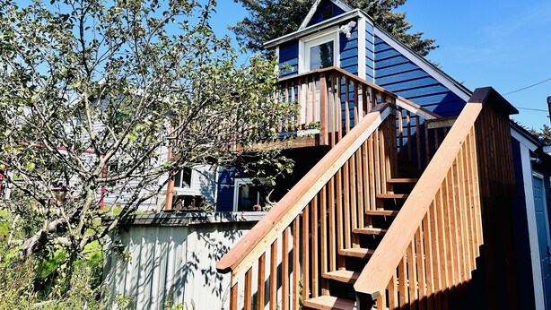 A wooden staircase leading to a blue-roofed upper level of a house with a view of trees and a clear sky.