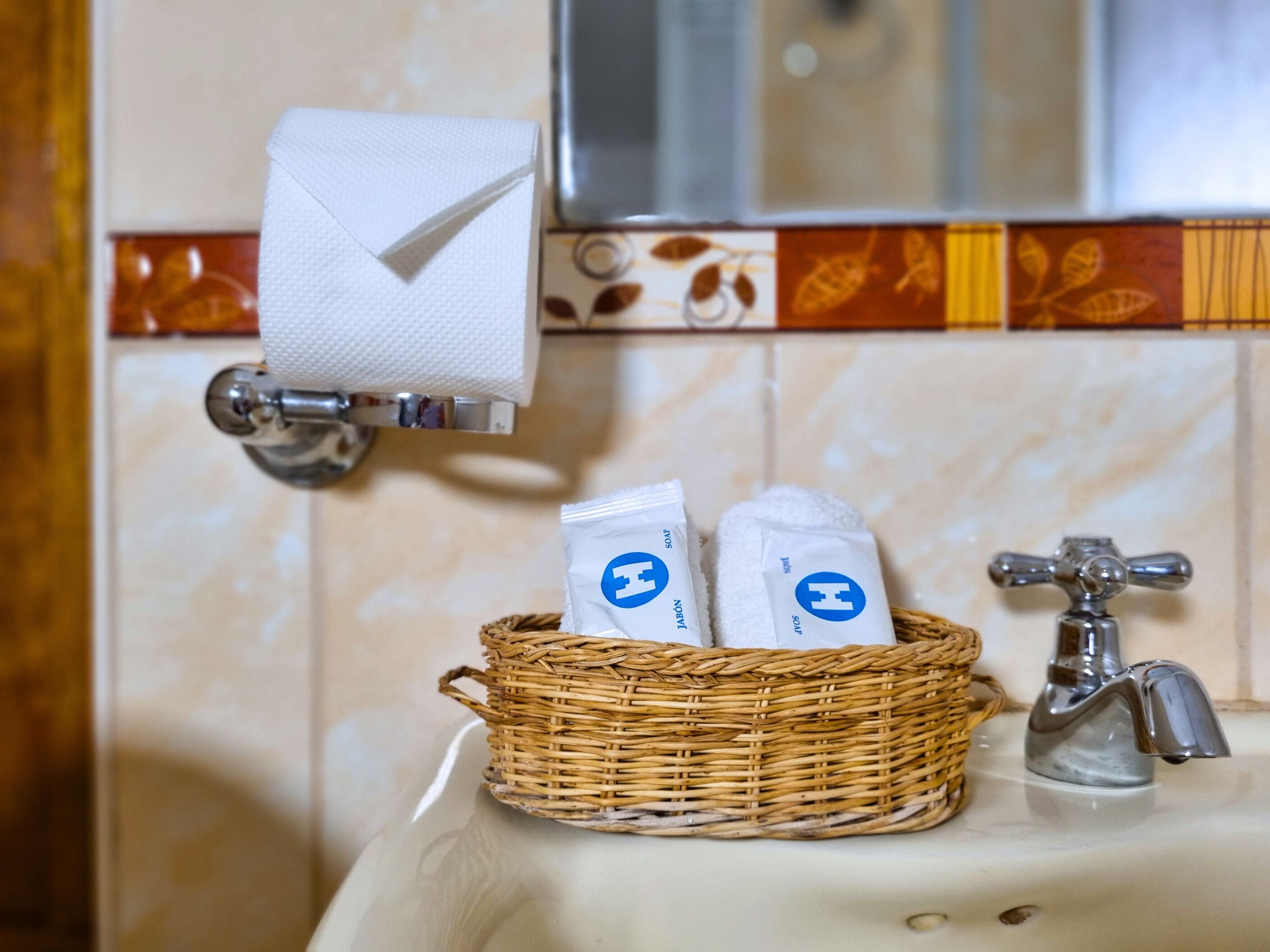 Et badeværelse med håndvask, spejl, håndklædedispenser og en fletkurv med toiletartikler.