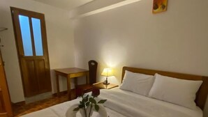 A hotel room with a neatly made bed, a wooden nightstand with a lamp, a small table, and a door with frosted glass panels. - Inka House Inn (Machu Picchu)