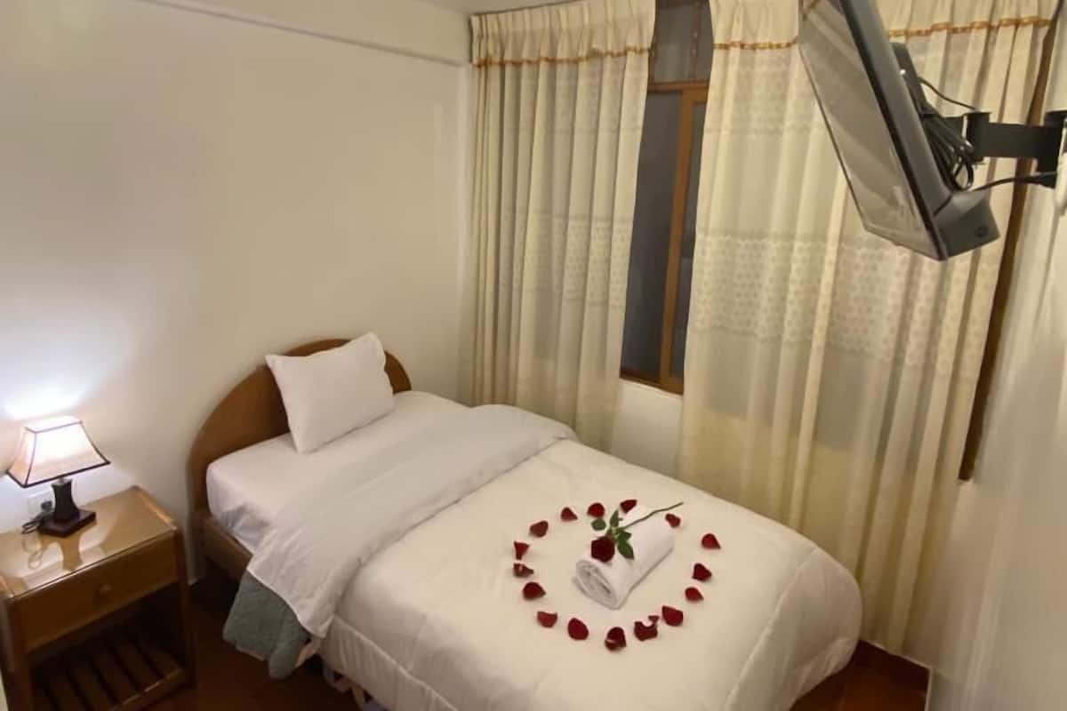 A neatly made bed with white linens, a bedside lamp, and a heart-shaped arrangement of rose petals on the bed.