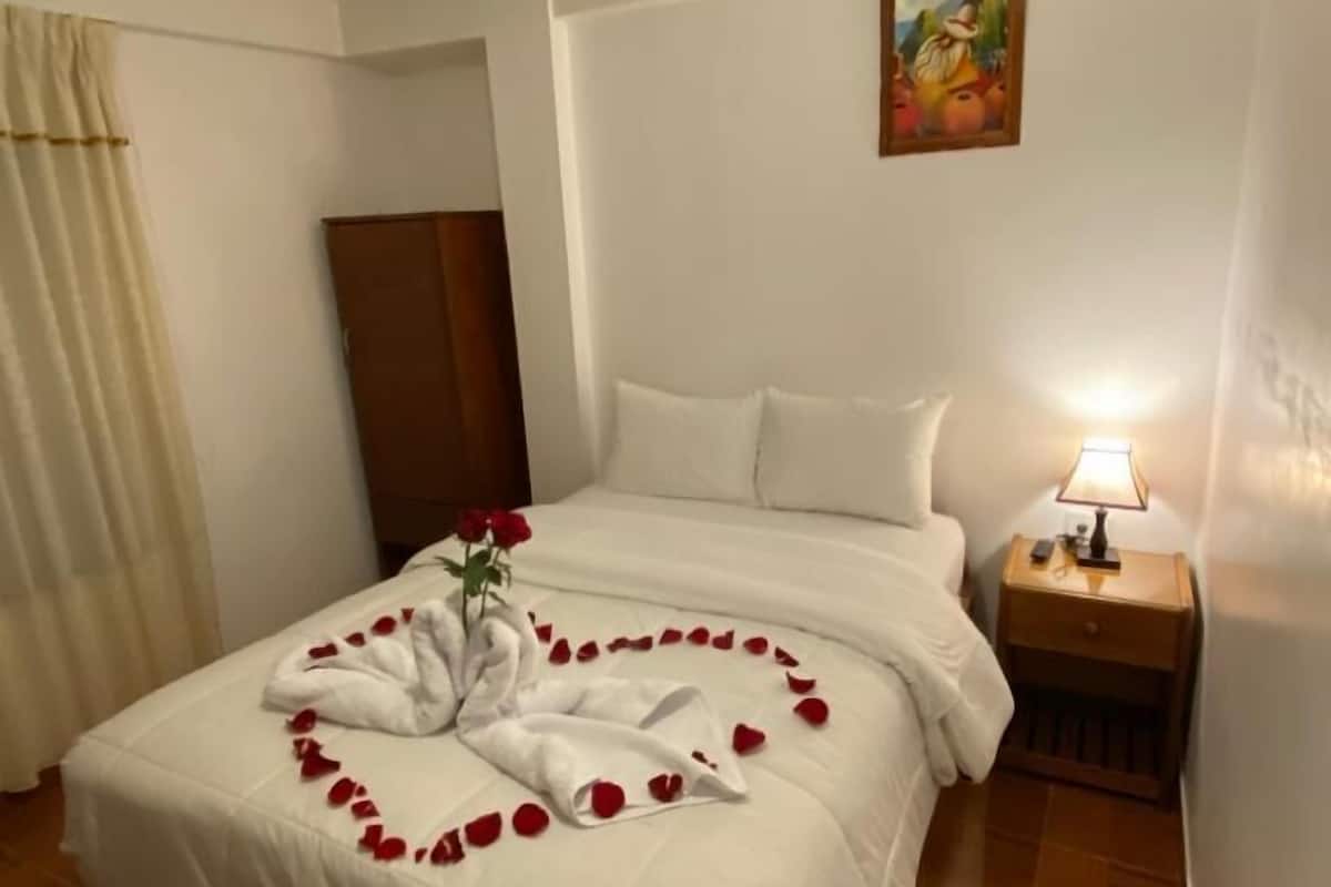 A neatly made bed with white linens, a heart-shaped arrangement of rose petals, and a bouquet of red roses.