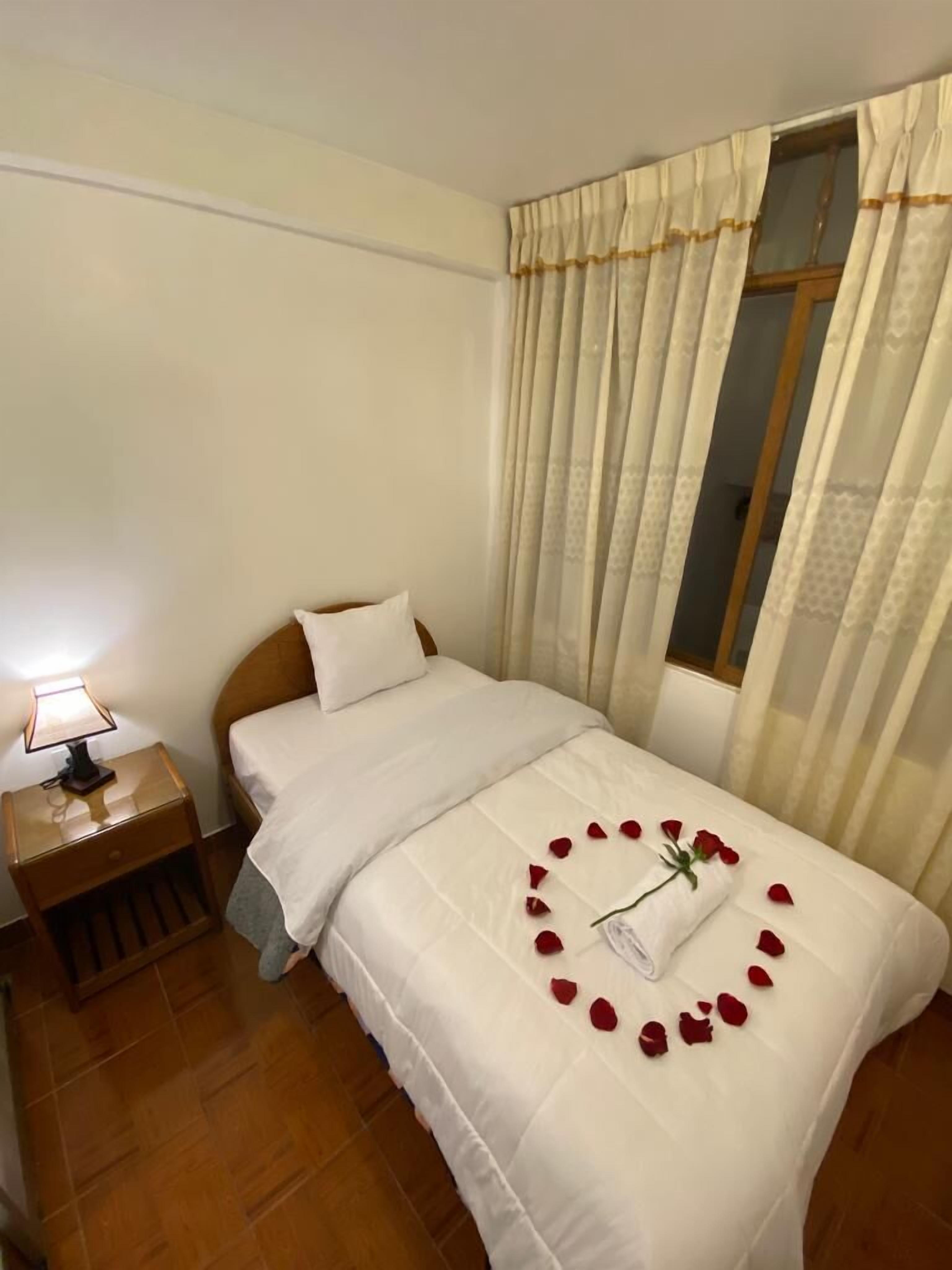 A neatly made bed with white bedding, a wooden nightstand with a lamp, and a window with sheer curtains.