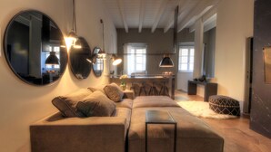 A modern living room with a sectional sofa, a small table, and a large mirror. There is a kitchen area in the background with bar stools and a countertop. - Luxury City Center Loft (Cagliari)