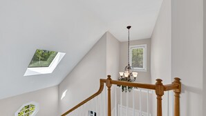 A staircase with a wooden handrail, white balusters, and a chandelier. There are two skylights on the ceiling, one with a view of trees and another with a view of a building.