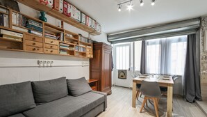 A modern living room with a grey sofa, wooden shelves with books, a dining table with chairs, and a window with curtains.