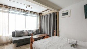 A bedroom with a bed, a wooden nightstand, a window with curtains, a sofa, and a small table.