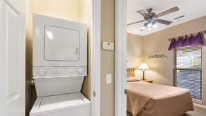 Une buanderie équipée d’un lave-linge et d’un sèche-linge à chargement frontal, d’un ventilateur de plafond, et une chambre adjacente avec un lit, une fenêtre et des rideaux violets.