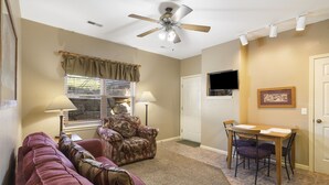 Un salon avec un ventilateur de plafond, un canapé, un fauteuil, une table à manger avec des chaises, une fenêtre avec des stores et une porte.