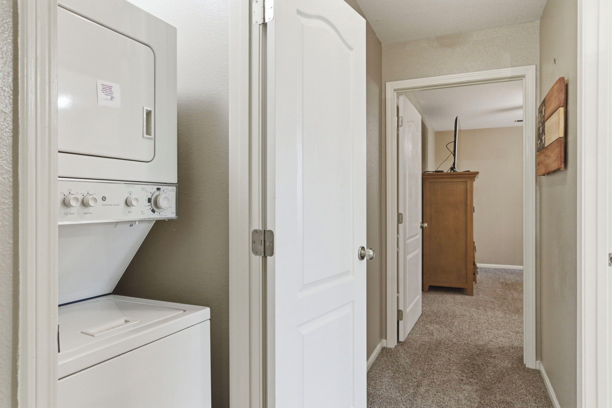 Un cuarto de lavandería con lavadora y secadora apiladas, un gabinete de madera y un pasillo que conduce a otra habitación.