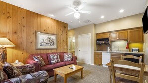 Ein Wohnzimmer mit Deckenventilator aus Holz, einem roten Sofa mit gemusterten Kissen, einem Holztisch und einer Küchenzeile mit Holzschränken und Geräten.