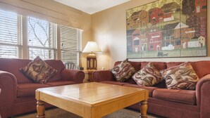 A living room with a wooden coffee table, two red sofas with patterned cushions, a floor lamp, and a large landscape painting on the wall.