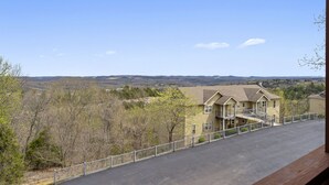 A two-story building with a driveway and a fenced area, surrounded by trees and hills in the background. - Relaxing Condo with Fireplace, Jetted Tub, and Private Balcony (Reeds Spring)