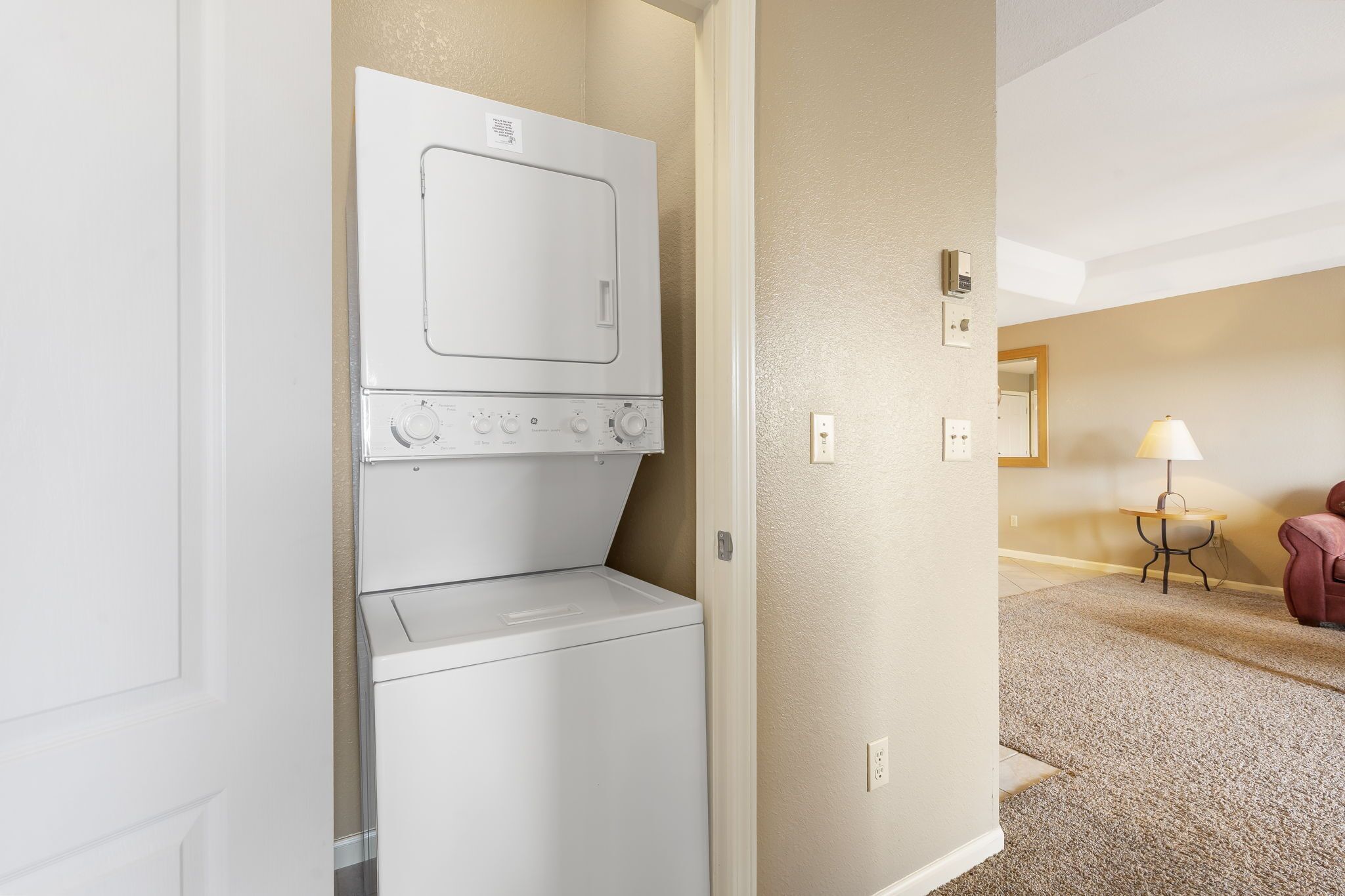 A laundry room with a stacked washer and dryer, beige walls, a small table with a lamp, and a red chair in the background.