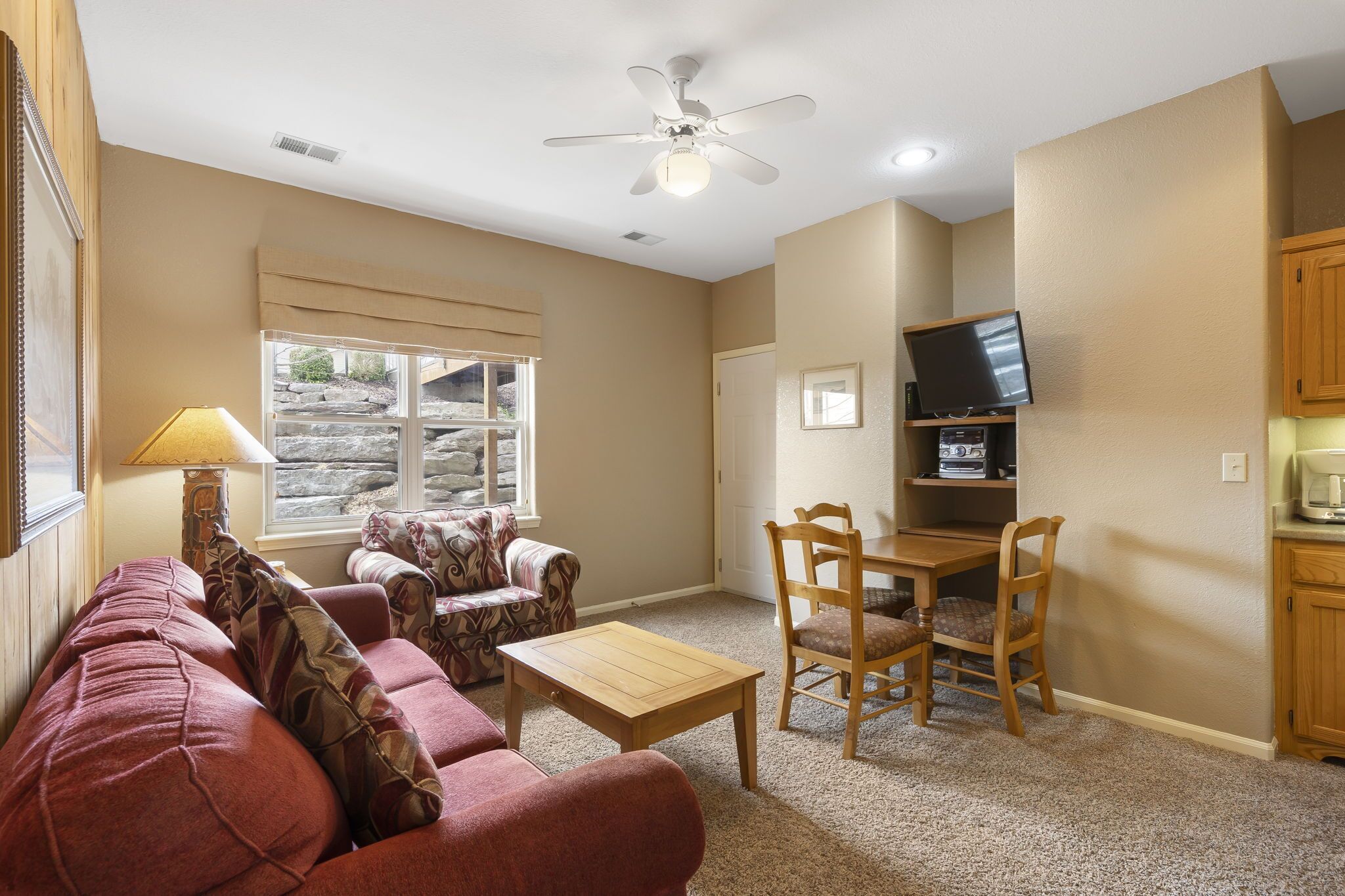A living room with a red couch, a wooden coffee table, a dining table with chairs, a ceiling fan, and a television on a wooden stand.