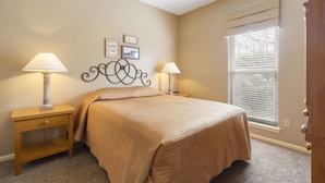 A bedroom with a bed, two bedside tables with lamps, a window with blinds, and a wall with framed pictures. - Quaint Condo Suite in a Peaceful Community near Silver Dollar City (Reeds Spring)