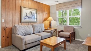 A living room with a wooden wall, a sofa, a chair, a coffee table, and a window with blinds. - Cute Queen Suite at StoneBridge  Village - minutes from SDC! (Reeds Spring)