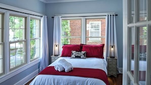 A bedroom with a large bed, two bedside tables, and a view of a brick building through the windows.