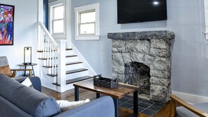 A living room with a stone fireplace, a flat-screen TV, a grey sofa, and a staircase with white railings.