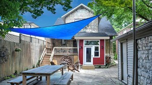 A stone patio with a wooden table and benches, a red house with a white door, and a blue shade sail.