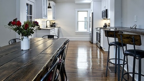 A modern kitchen with white cabinetry, a wooden dining table, and a view to the living area.