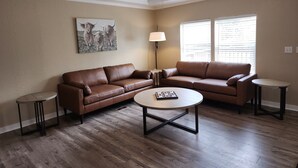 A living room with a brown leather sectional sofa, a round coffee table, and two side tables. There is a ceiling fan, a floor lamp, and a painting on the wall. - Cattlemens Quarters beautiful ranch setting with fenced yards and a great deck! (Tyler)
